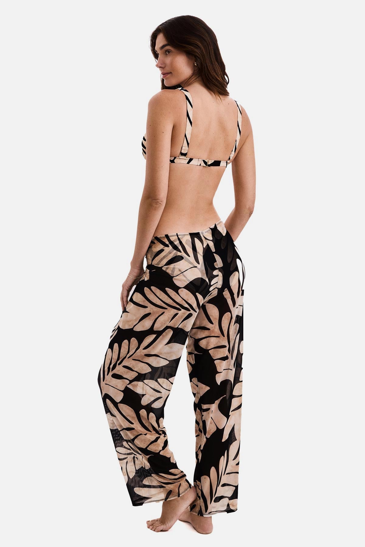 Woman turned left wearing black and tan palm leaf printed swim cover up pants with a matching bikini top